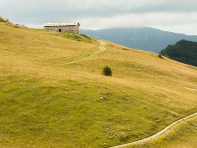 Rifugio in pietra sull’altopiano della Lessinia, circondato da prati dorati e raggiunto da un sentiero sterrato, con montagne sullo sfondo